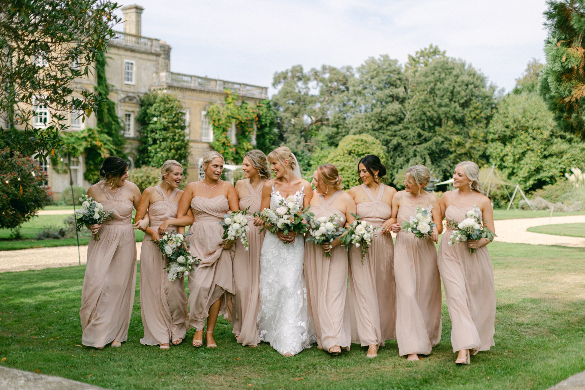 Bridesmaid wearing luxury blush dresses laughing at each other in the grounds of a stately home on a sunny day